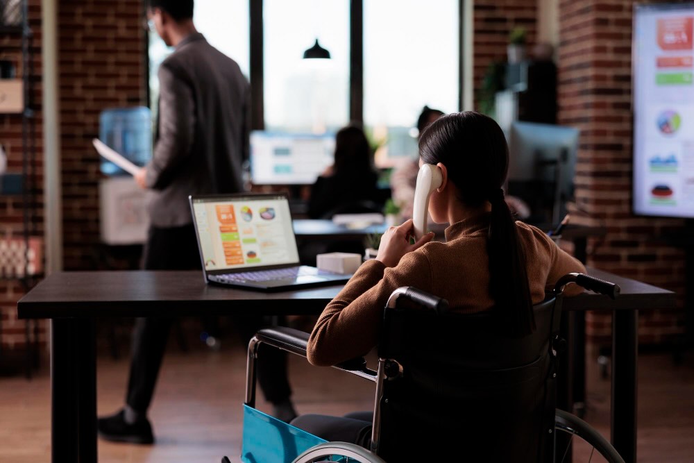 DE&I Courses for Managers- Your First Step to a More Professional, Inclusive Workplace DE&I Courses for Managers - Person in a wheelchair working in an office environment, using a telephone and viewing a presentation on a laptop screen